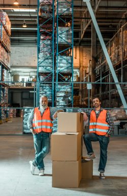 male-warehouse-workers-in-vests-standing-with-boxes-and-smiling-at-camera.jpg